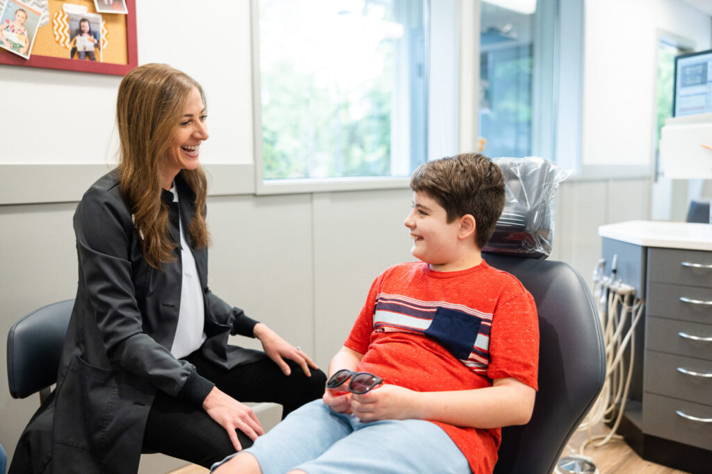 dentist smiling and talking with a young boy sitting in a dental chair, holding sunglasses in a bright and modern dental office