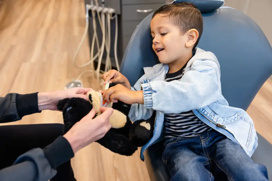 A young boy practices brushing teeth on a black bear stuff toy